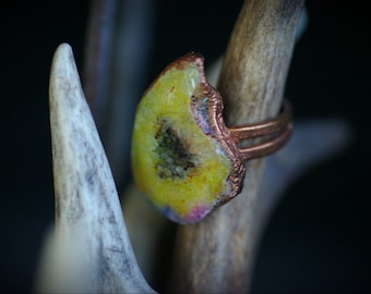 May include: A copper ring with a yellow, green, and pink agate stone set in the band. The ring is displayed on a piece of antler.