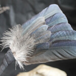 Puede incluir: Un abanico de plumas azul y gris con un mango de cinta negra. Las plumas est&aacute;n dispuestas en forma de abanico y est&aacute;n atadas con una cinta negra.
