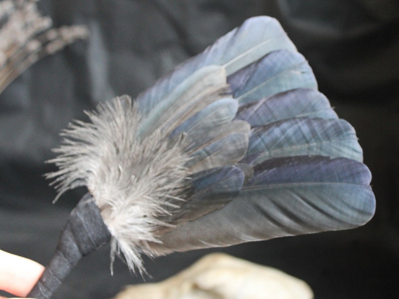 Puede incluir: Un abanico de plumas azul y gris con un mango de cinta negra. Las plumas est&aacute;n dispuestas en forma de abanico y est&aacute;n atadas con una cinta negra.