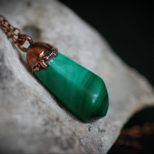 Puede incluir: Un colgante de cobre con una piedra de malaquita verde. La piedra es larga y puntiaguda, con un patrón natural estriado. El cobre está oxidado y tiene un color marrón oscuro.