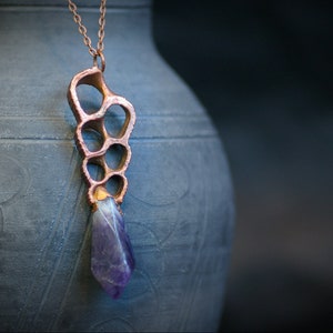 Puede incluir: Un collar de cobre con un colgante geométrico que presenta un grupo de círculos abiertos y una piedra de amatista púrpura.