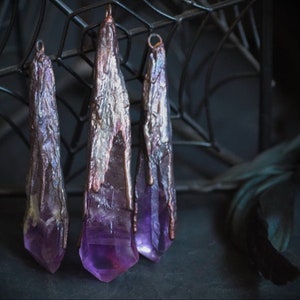 Puede incluir: Tres colgantes de cristal de amatista con un acabado de cobre. Los cristales son largos y puntiagudos, y el acabado de cobre es texturizado y tiene un brillo metálico.
