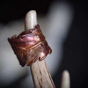 May include: A copper ring with a textured, leaf-like design. The ring is displayed on a piece of antler.