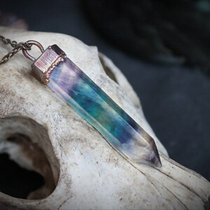 Puede incluir: Un collar de cadena de cobre con un colgante que presenta un cristal largo, facetado e iridiscente con tonos de morado, azul y verde.