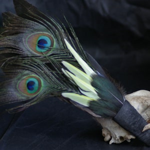Puede incluir: Un abanico de plumas de pavo real con plumas verdes, azules y amarillas unidas a un cráneo negro con una banda negra.