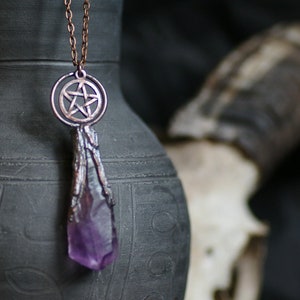 Puede incluir: Un collar de cobre con un colgante de pentagrama y una gran punta de cristal de amatista. El collar está colgado sobre un fondo oscuro.