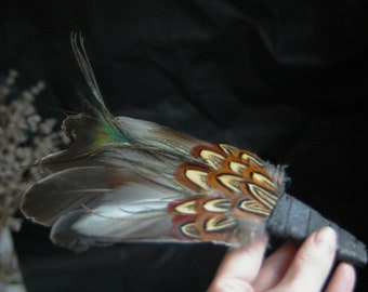 May include: A hand fan made of pheasant feathers with a black leather handle. The feathers are brown, white, and orange with green and blue accents.
