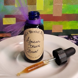 May include: A small, dark blue glass bottle with a purple ribbon and a label that reads "African Dream Bean." A glass dropper with a black top sits next to the bottle on a white plate. The background features an abstract painting.
