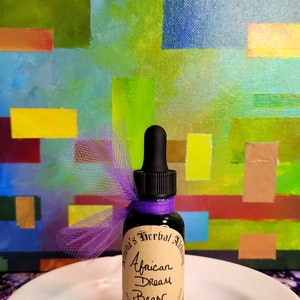 May include: A small, dark glass bottle with a dropper, labeled "African Dream Bean" and "Athena's Herbal Alchemy." The bottle is adorned with a purple ribbon and sits on a white plate. The background features an abstract painting with colorful blocks.
