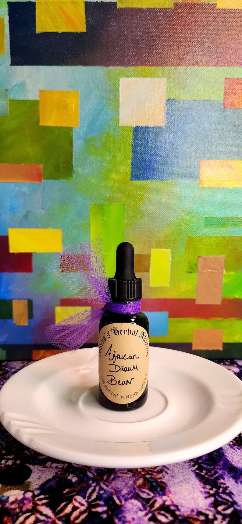 May include: A small, dark glass bottle with a dropper, labeled "African Dream Bean" and "Athena's Herbal Alchemy." The bottle is adorned with a purple ribbon and sits on a white plate. The background features an abstract painting with colorful blocks.