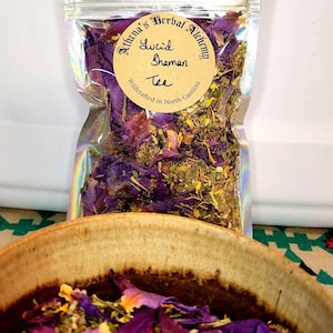 May include: A clear, iridescent bag filled with dried herbs and flowers, labeled "Lucid Shaman Tee" from Athena's Herbal Alchemy, wildcrafted in North Carolina. A ceramic bowl with dried herbs and purple petals is in the foreground.