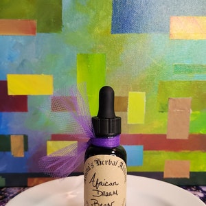 May include: A small, dark glass bottle with a black dropper, labeled "African Dream Bean" and tied with purple ribbon and tulle. The bottle sits on a white plate against a colorful abstract background.