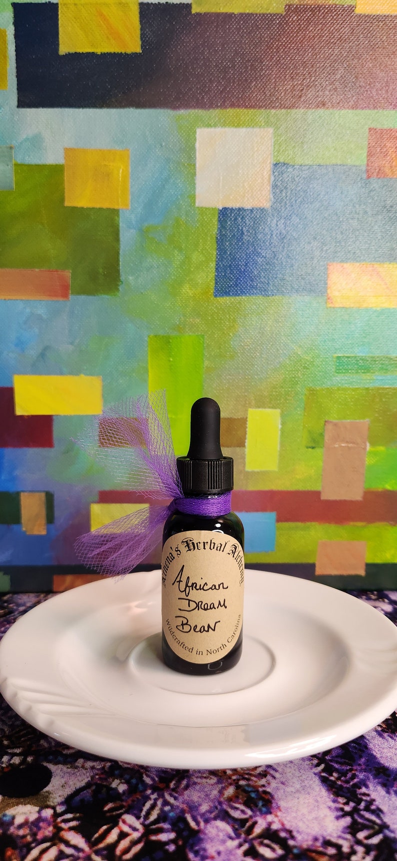 May include: A small, dark glass bottle with a black dropper, labeled "African Dream Bean" and tied with purple ribbon and tulle. The bottle sits on a white plate against a colorful abstract background.