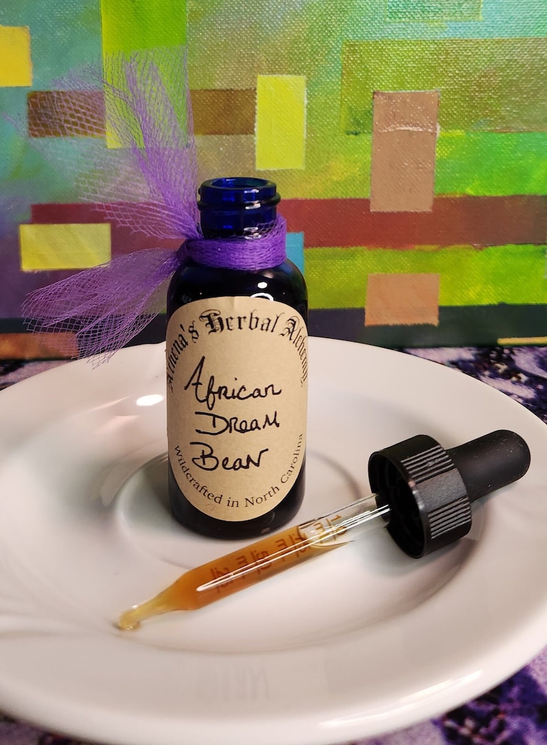 May include: A small, dark blue glass bottle with a purple ribbon and a label that reads "African Dream Bean." A glass dropper with a black top sits next to the bottle on a white plate. The background features an abstract painting.