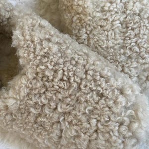 May include: A pair of slip-on slippers crafted from a thick, cream-coloured faux shearling material. The exterior features a dense, curly, and textured pile that covers the entire upper surface of the footwear, providing a soft and uniform appearance.