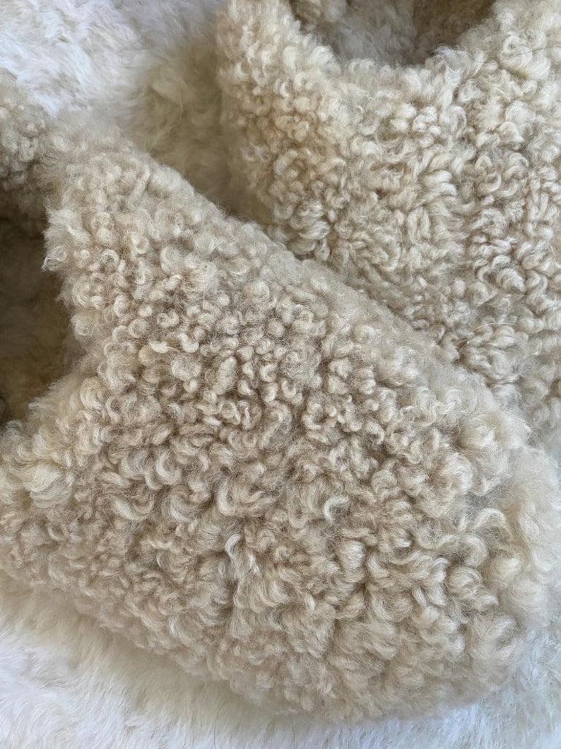 May include: A pair of slip-on slippers crafted from a thick, cream-coloured faux shearling material. The exterior features a dense, curly, and textured pile that covers the entire upper surface of the footwear, providing a soft and uniform appearance.