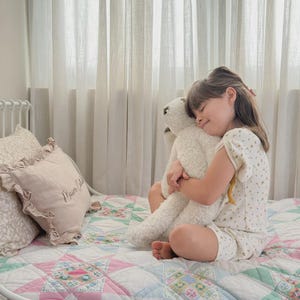 May include: A cream-colored plush teddy bear with a textured, curly fleece-like surface. The bear has a rounded head, small ears, and long limbs, and is held in a close embrace. The toy is constructed from soft, fuzzy material with visible stitching.