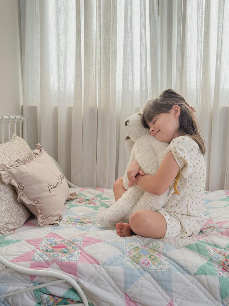 May include: A cream-colored plush teddy bear with a textured, curly fleece-like surface. The bear has a rounded head, small ears, and long limbs, and is held in a close embrace. The toy is constructed from soft, fuzzy material with visible stitching.