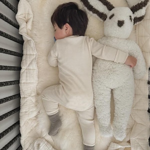 May include: A child wearing a cream-colored waffle-knit long-sleeve shirt and matching pants with grey socks, sleeping next to a large, plush white rabbit stuffed animal with dark brown textured ears and facial features on a soft cream blanket.
