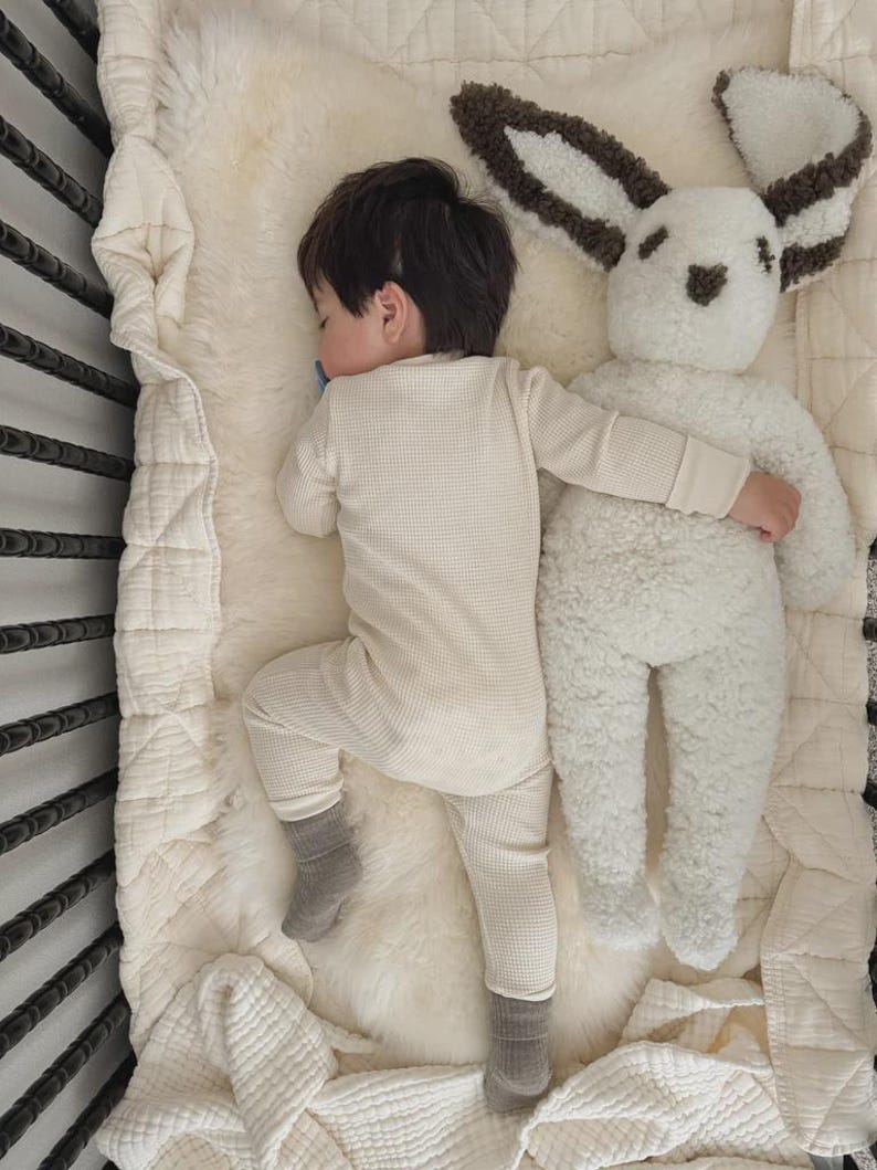 May include: A child wearing a cream-colored waffle-knit long-sleeve shirt and matching pants with grey socks, sleeping next to a large, plush white rabbit stuffed animal with dark brown textured ears and facial features on a soft cream blanket.