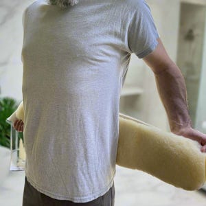 May include: A light gray t-shirt and dark gray pants. A person is holding a large, cylindrical, light yellow paint roller. The paint roller has a fluffy texture and is ready for use.