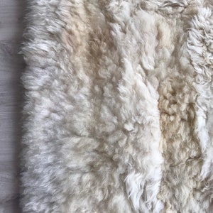 Pode incluir: Um tapete de pele de carneiro branco com uma textura macia e fofa. O tapete &eacute; feito de pele de carneiro natural e tem uma cor natural branco-queimado.