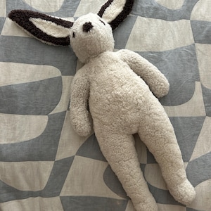 May include: A plush rabbit toy made of cream-colored, textured sherpa fabric. The rabbit features long, upright ears with dark brown edges and a matching dark brown nose. The body is soft and rounded, with simple limbs and a neutral, fuzzy appearance.