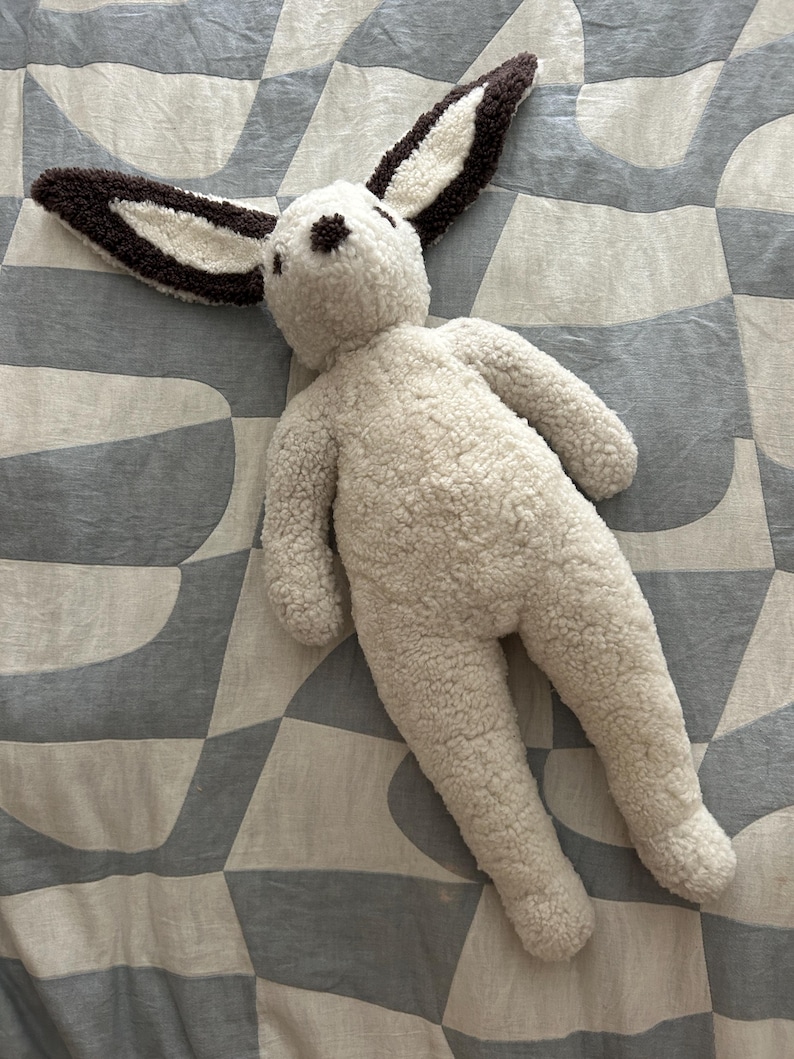 May include: A plush rabbit toy made of cream-colored, textured sherpa fabric. The rabbit features long, upright ears with dark brown edges and a matching dark brown nose. The body is soft and rounded, with simple limbs and a neutral, fuzzy appearance.