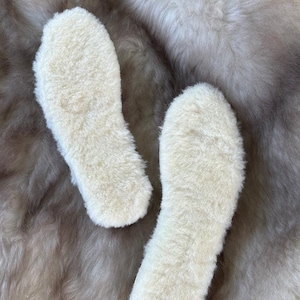 May include: A pair of off-white, fluffy sheepskin insoles with a thick, textured pile surface. The insoles are shaped to fit the contours of a foot, featuring a rounded heel and a tapered toe area, showcasing the dense, soft wool fibres.