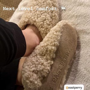 May include: Beige shearling slippers with a textured, curly wool exterior and a tan suede sole. The sole features the embossed text "EAST PERRY" and the size "39". The text "Next level comfort" and a sheep icon are visible on the upper portion.
