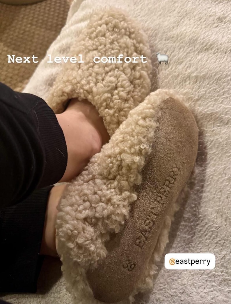 May include: Beige shearling slippers with a textured, curly wool exterior and a tan suede sole. The sole features the embossed text "EAST PERRY" and the size "39". The text "Next level comfort" and a sheep icon are visible on the upper portion.