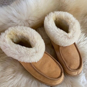 Puede incluir: Un par de zapatillas de ante marr&oacute;n con forro y ribete de piel sint&eacute;tica blanca. Las zapatillas est&aacute;n sobre una superficie blanca y esponjosa.