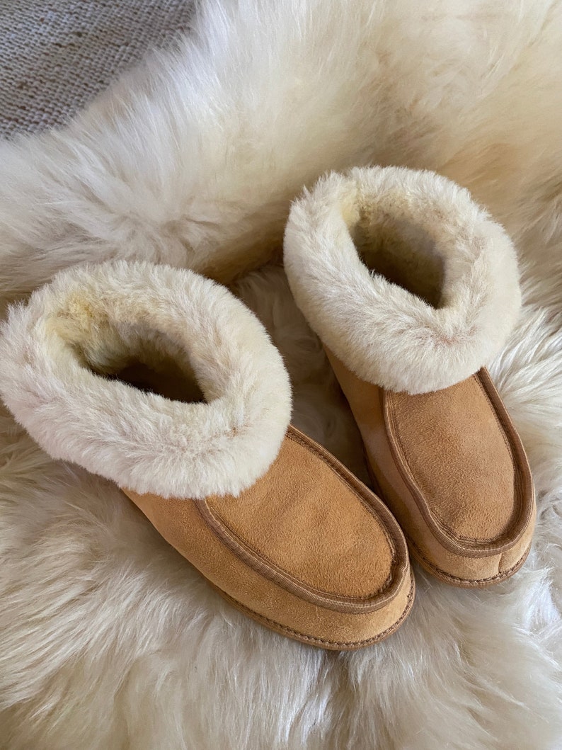 Puede incluir: Un par de zapatillas de ante marr&oacute;n con forro y ribete de piel sint&eacute;tica blanca. Las zapatillas est&aacute;n sobre una superficie blanca y esponjosa.