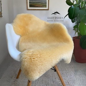 May include: A soft, pale yellow sheepskin rug draped over a white molded plastic chair with wooden legs. The wall behind features the text EAST PERRY NATURE. IN THE HOME with a mountain graphic. The rug has a thick, dense, and plush texture.