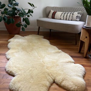 May include: A cream-colored sheepskin rug with a long, rectangular shape. The rug is laid out on a hardwood floor in a living room setting.