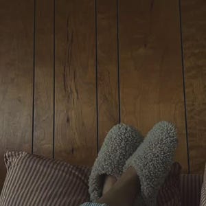 May include: A pair of light grey, fuzzy, sherpa-style slip-on slippers with a rounded toe design. The slippers feature a thick, textured fleece material and are worn over light blue and white striped fabric, resting against a red and white striped pillow.