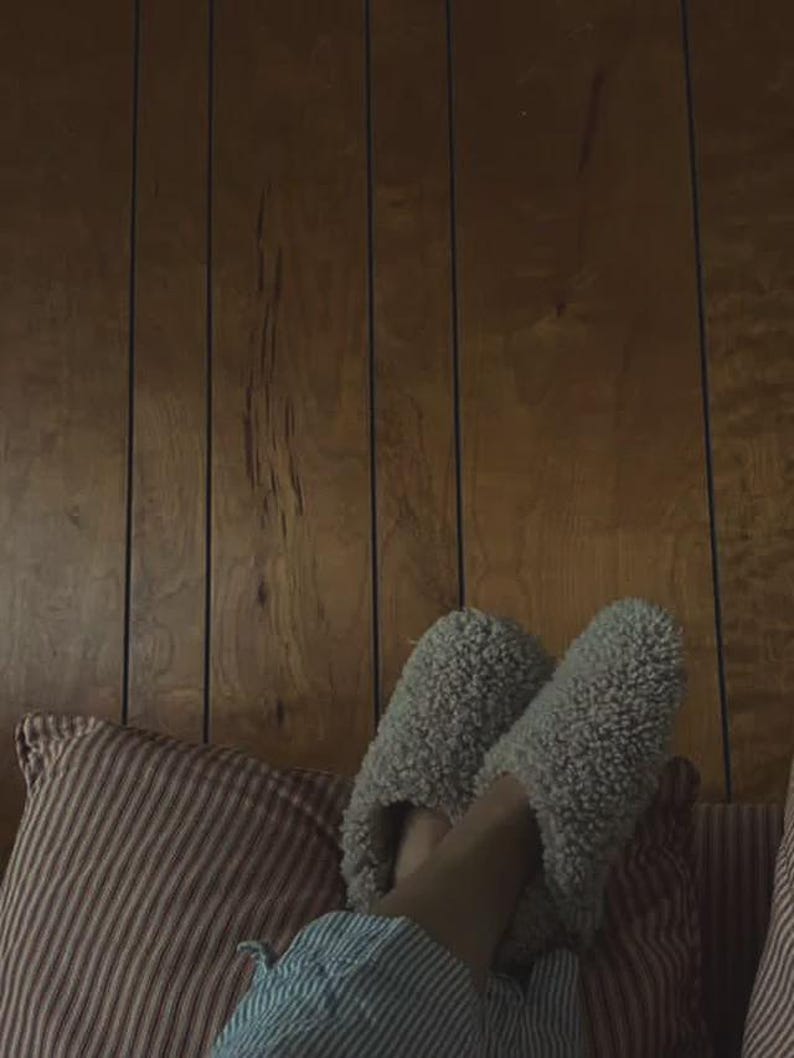 May include: A pair of light grey, fuzzy, sherpa-style slip-on slippers with a rounded toe design. The slippers feature a thick, textured fleece material and are worn over light blue and white striped fabric, resting against a red and white striped pillow.