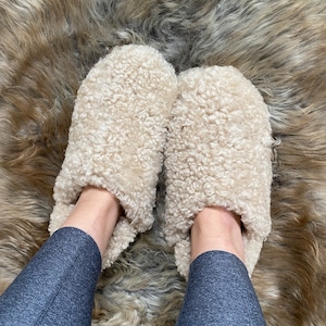 May include: A pair of cream-colored, slip-on slippers featuring a thick, curly, textured fleece-like exterior. The slippers have a rounded toe design and a plush, soft appearance, providing full coverage for the top of the foot in a neutral tone.