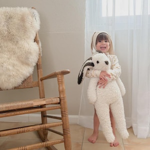 May include: A large, cream-colored plush rabbit toy with long, dark brown and white ears. The stuffed animal features a soft, textured fabric body, a small dark nose, and dark eyes. It is held by a child wearing a matching cream-colored bunny ear hat.