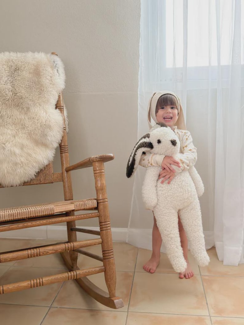 May include: A large, cream-colored plush rabbit toy with long, dark brown and white ears. The stuffed animal features a soft, textured fabric body, a small dark nose, and dark eyes. It is held by a child wearing a matching cream-colored bunny ear hat.