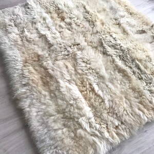 Pode incluir: Um tapete de pele de carneiro branco com uma textura macia e fofa. O tapete &eacute; feito de pele de carneiro natural e tem uma cor bege claro.