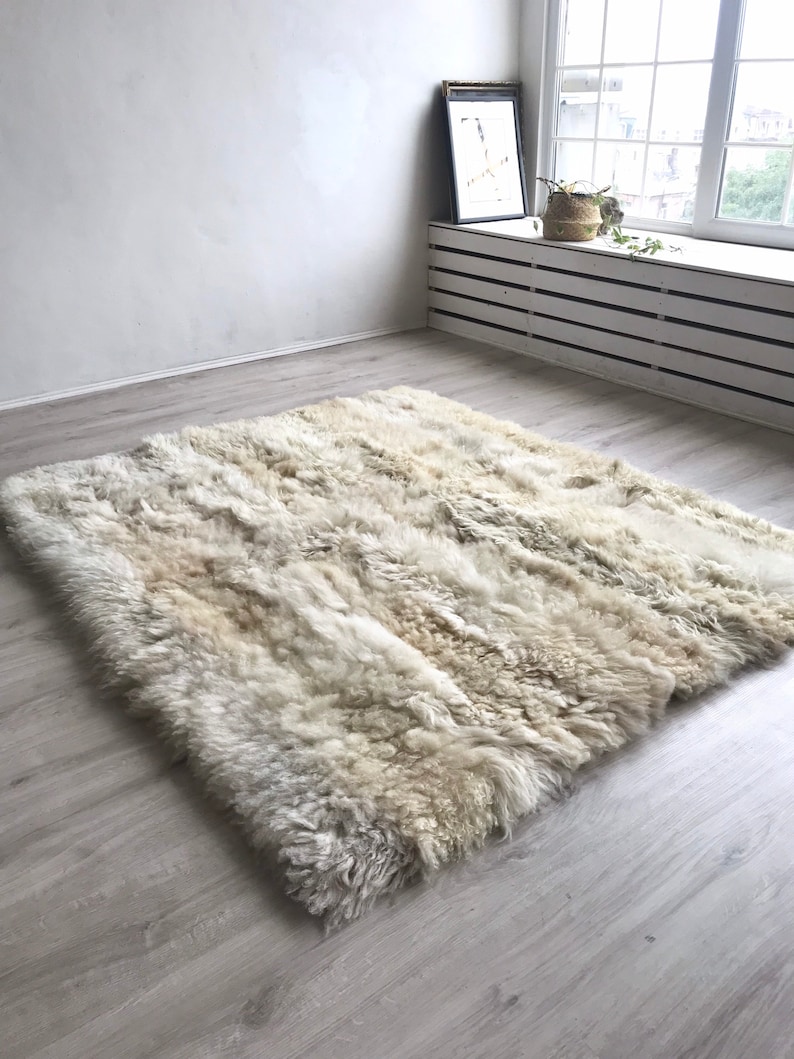 Pode incluir: Um tapete de pele de carneiro branco e felpudo com uma cor natural, branco quebrado. O tapete est&aacute; colocado sobre um piso de madeira clara em uma sala com uma janela e uma parede branca.