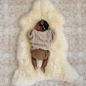 May include: A baby wearing a brown sweater and brown pants is sleeping on a white sheepskin rug. The rug is on a patterned floor.