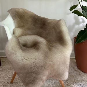 May include: A sheepskin rug in shades of beige and brown is draped over a white chair with wooden legs. The rug's texture is soft and fluffy, creating a cozy and inviting look. The chair is set against a neutral background.