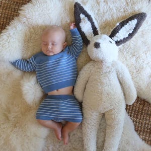 May include: A sleeping infant wearing a blue long-sleeved shirt and matching shorts with thin white horizontal stripes, positioned next to a large, plush, cream-colored stuffed rabbit toy with dark brown and white ears and a dark brown nose.