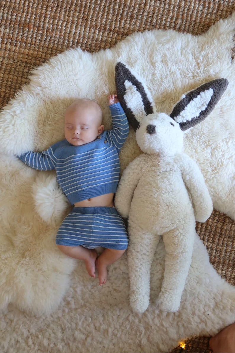May include: A sleeping infant wearing a blue long-sleeved shirt and matching shorts with thin white horizontal stripes, positioned next to a large, plush, cream-colored stuffed rabbit toy with dark brown and white ears and a dark brown nose.