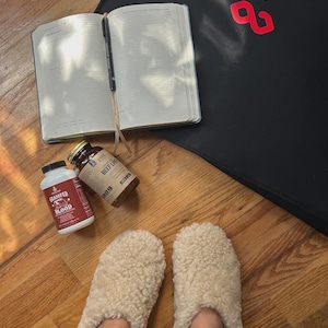 May include: A pair of cream-coloured fuzzy sherpa slippers, an open planner with a black pen, two supplement bottles labelled Grassfed Blood and Beef Liver, and a black mat with the red logo text BON CHA visible on the surface.