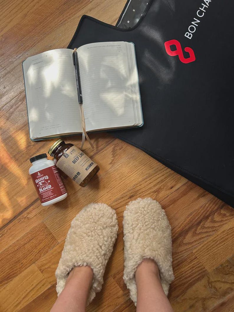 May include: A pair of cream-coloured fuzzy sherpa slippers, an open planner with a black pen, two supplement bottles labelled Grassfed Blood and Beef Liver, and a black mat with the red logo text BON CHA visible on the surface.