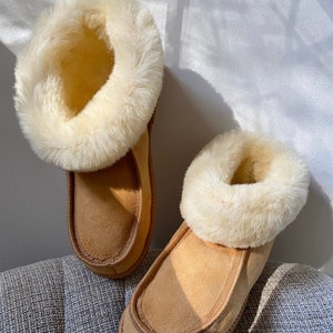 Puede incluir: Un par de zapatillas de ante marr&oacute;n con un ribete de piel sint&eacute;tica blanca. Las zapatillas est&aacute;n sobre una superficie gris texturizada.