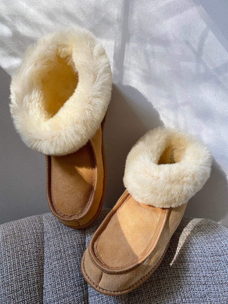 Puede incluir: Un par de zapatillas de ante marr&oacute;n con un ribete de piel sint&eacute;tica blanca. Las zapatillas est&aacute;n sobre una superficie gris texturizada.